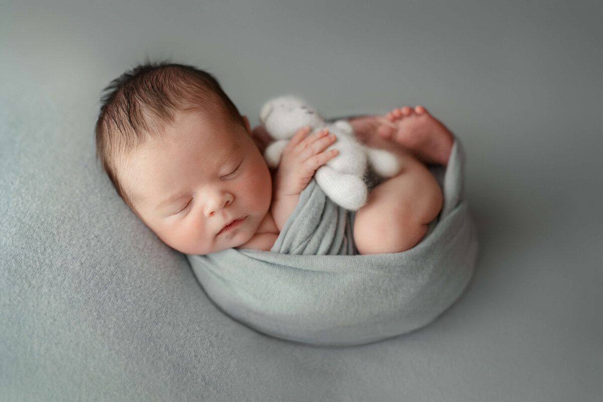 newborn baby boy in a light blue swaddle holding a bear on a light blue backdrop at his newborn photography session  with a photographer in a sterling va photo studio