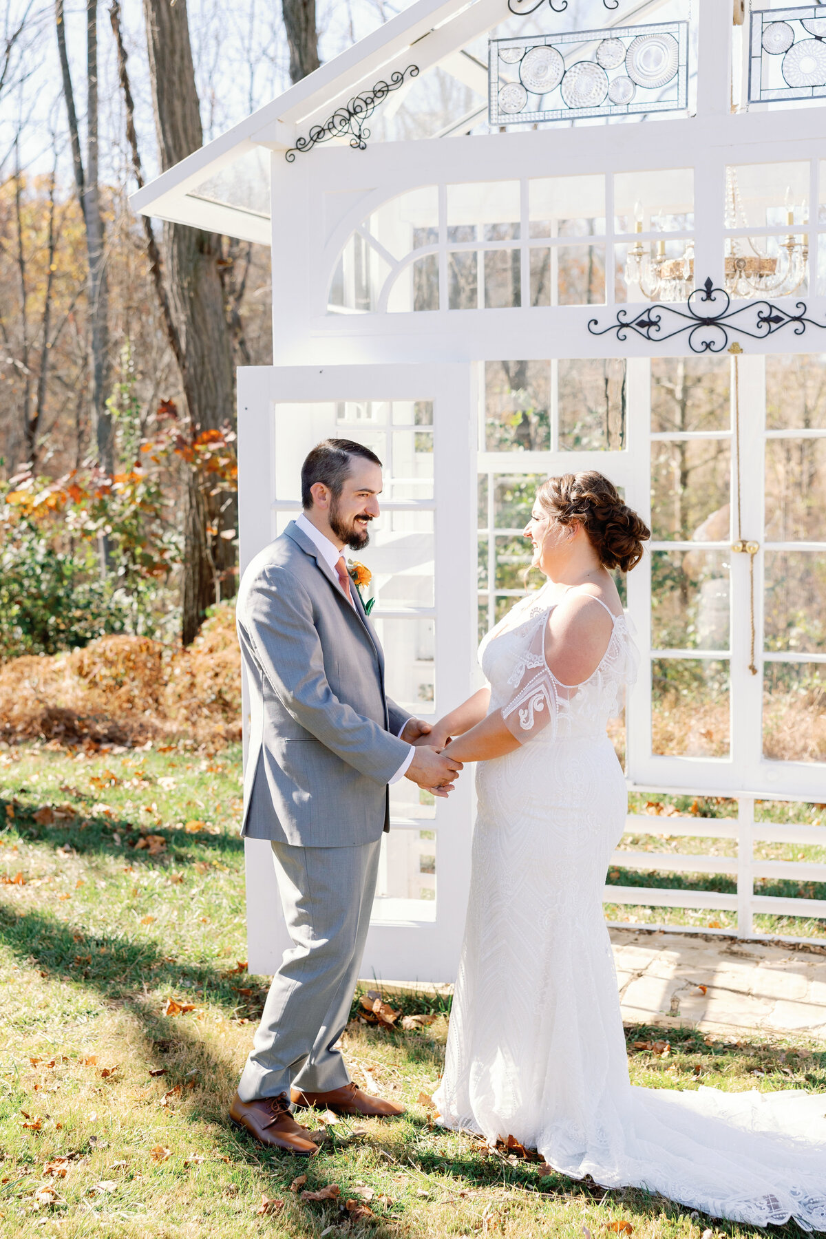 Groom in gray suit and bride in lace gown holding hands beneath white arbor during first look at Maryland wedding venue.