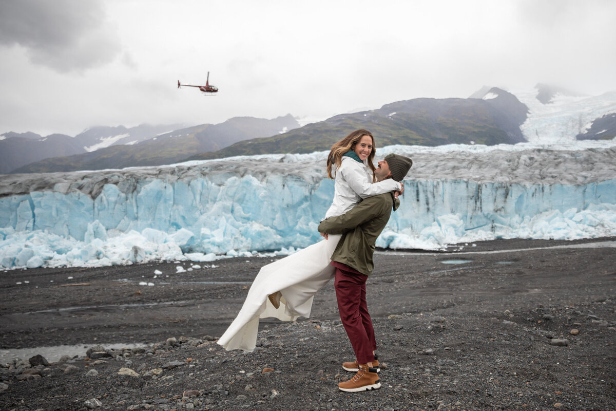 A groom picks his bride up as a helicopter flies overhead.