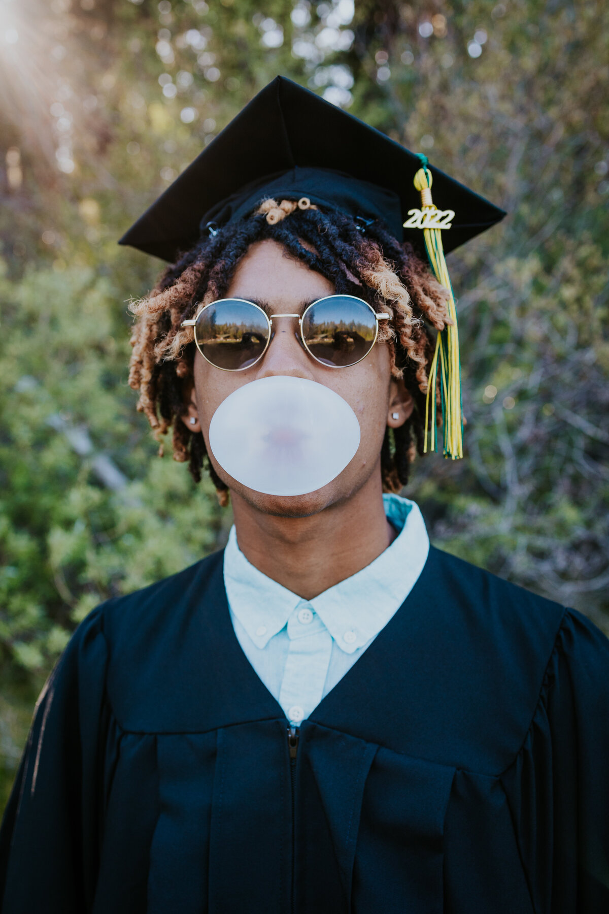 Young man wearing sunglasses and cap and gown blows giant bubble from gum.