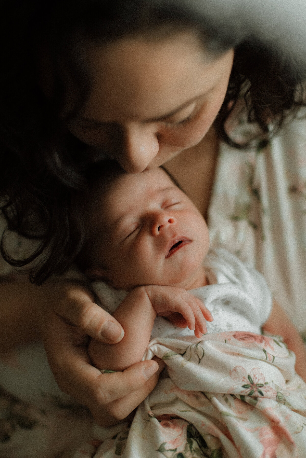 Mother gently kissing her newborn’s forehead while holding them close, wrapped in a floral swaddle, in soft natural light.