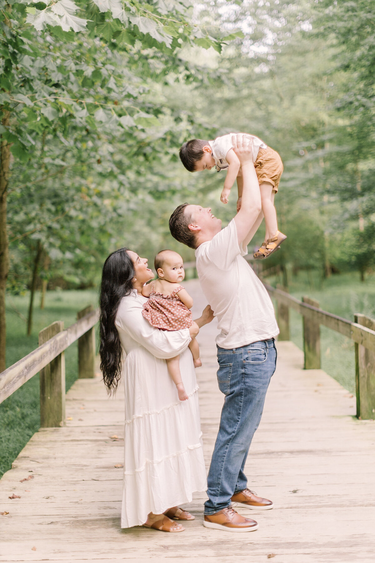 A joyful family standing on a wooden bridge surrounded by greenery, parents lifting their children in the air — Raleigh portrait photos.