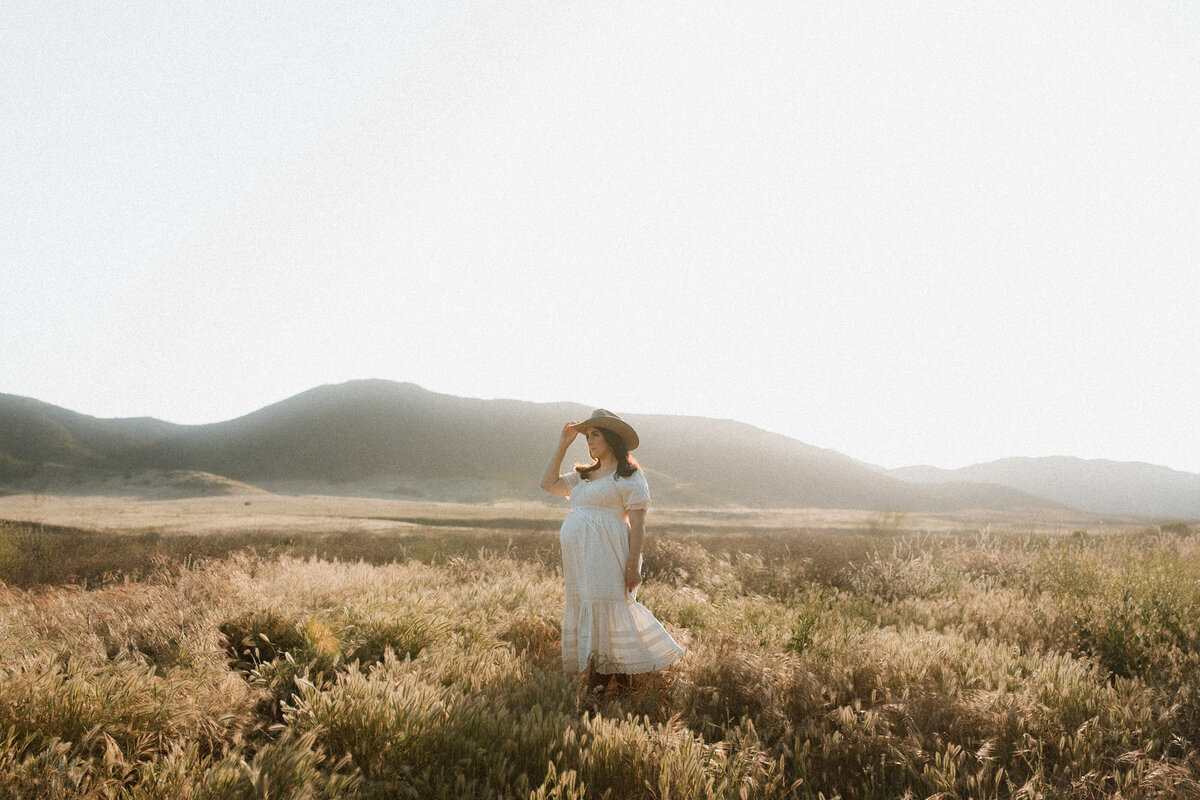 A pregnant woman in a white dress and cowboy hat stands in a sunlit field, holding the brim of her hat as soft light spills over the hills behind her during a maternity session in Temecula.