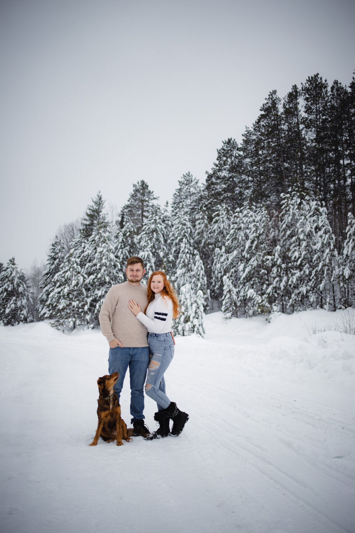 A couple stands smiling at the camera while their dog looks up at them surrounded by snow.