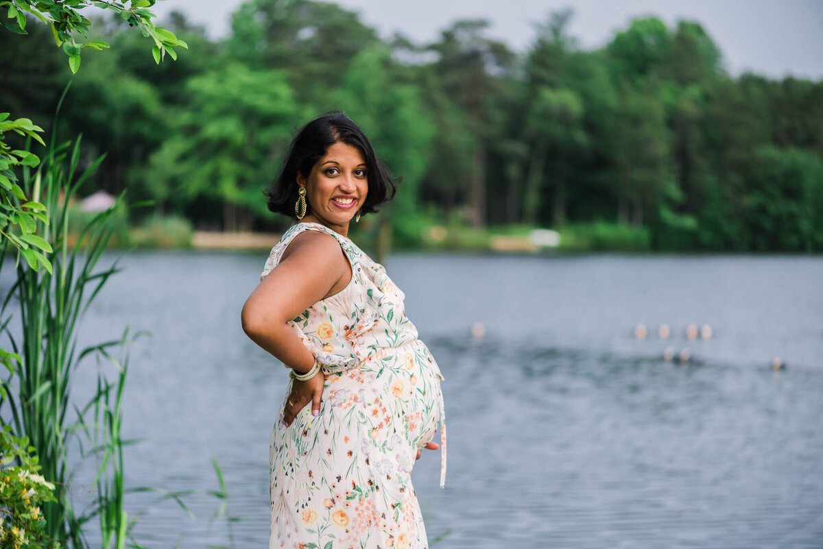 A glowing mom in a floral dress standing by a lake by Denise Van Photography