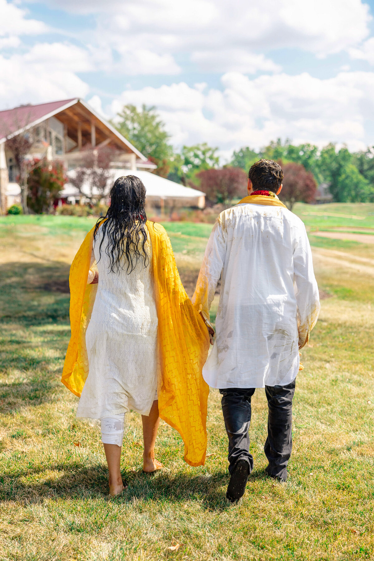 A couple walks hand in hand away from the camera across a grassy field. They wear traditional white and yellow attire. A large house and trees are visible in the background under a partly cloudy sky.