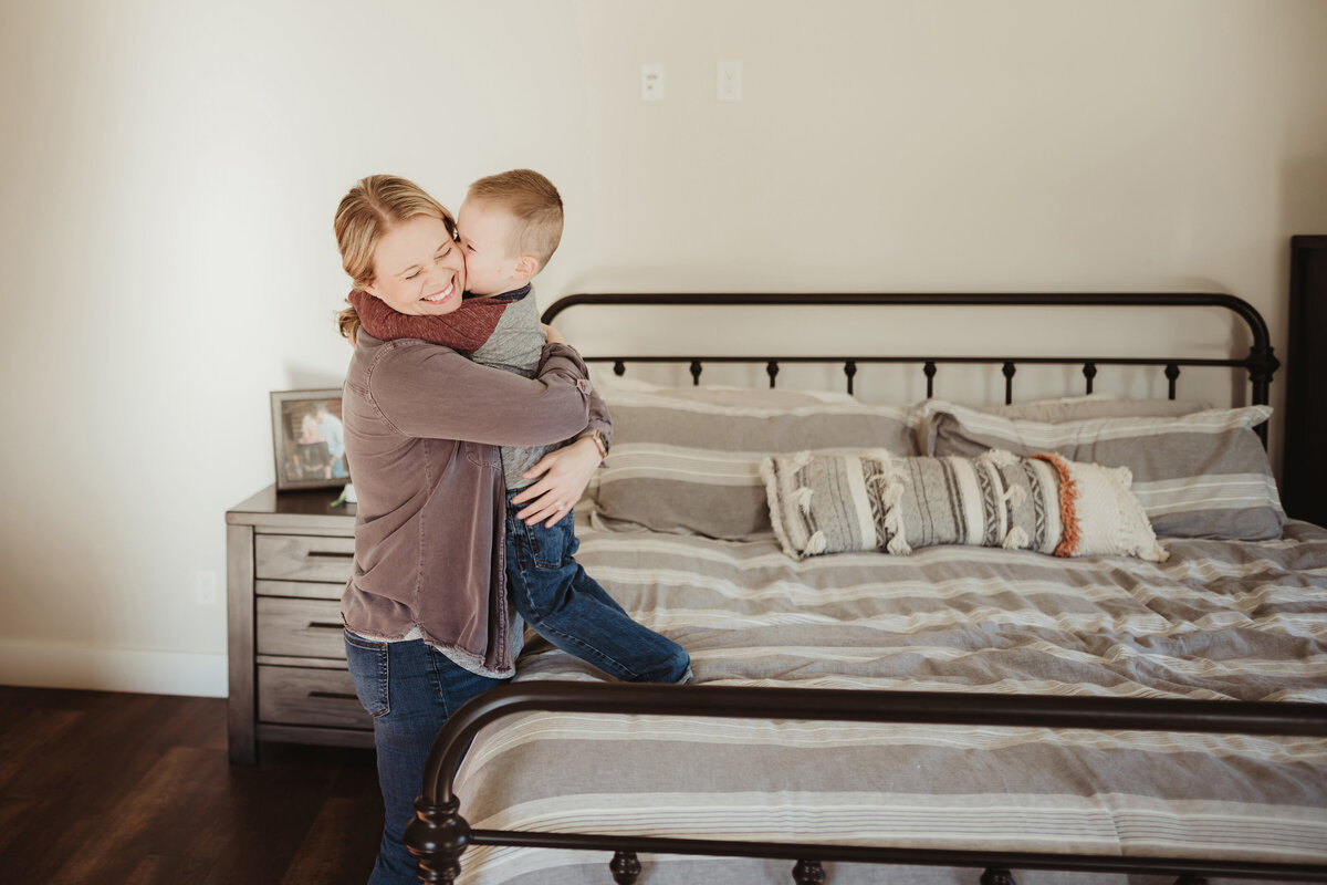A little boy is standing on the bed kissing and hugging his mom.
