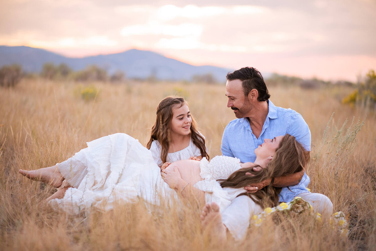 A family of three sit in the brown grass together. Dad embraces his pregnant wife who is laying in his lap an d young daughter sits next to then smiling at them. Taken by Brittany Danielle Photography