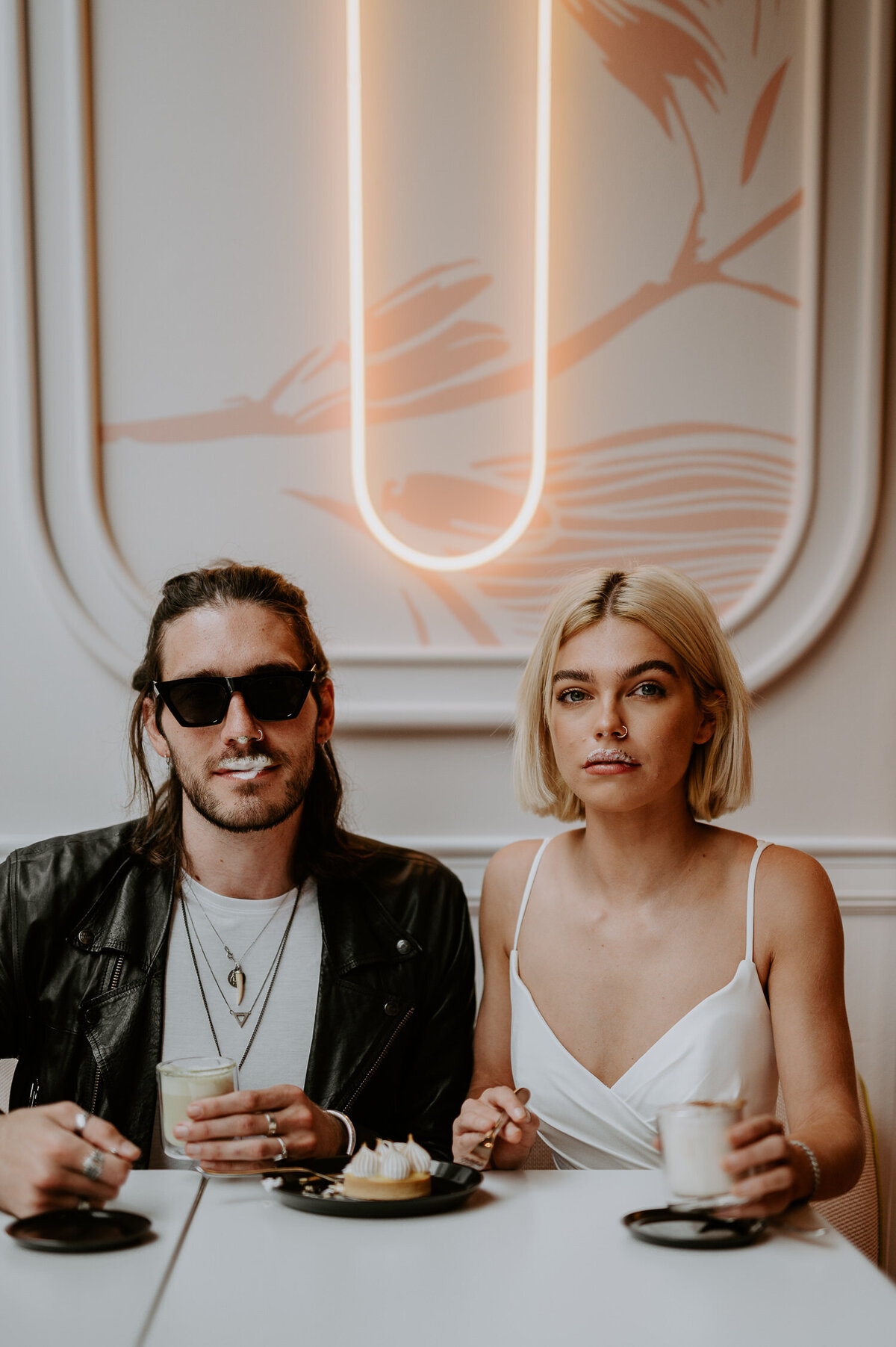 A couple sit in a coffee shop in Soho, London. They both have coffee moustaches, the groom has glasses, long hair and is wearing a leather jacket. The bride is wearing a strappy wedding dress from Justin Alexander.