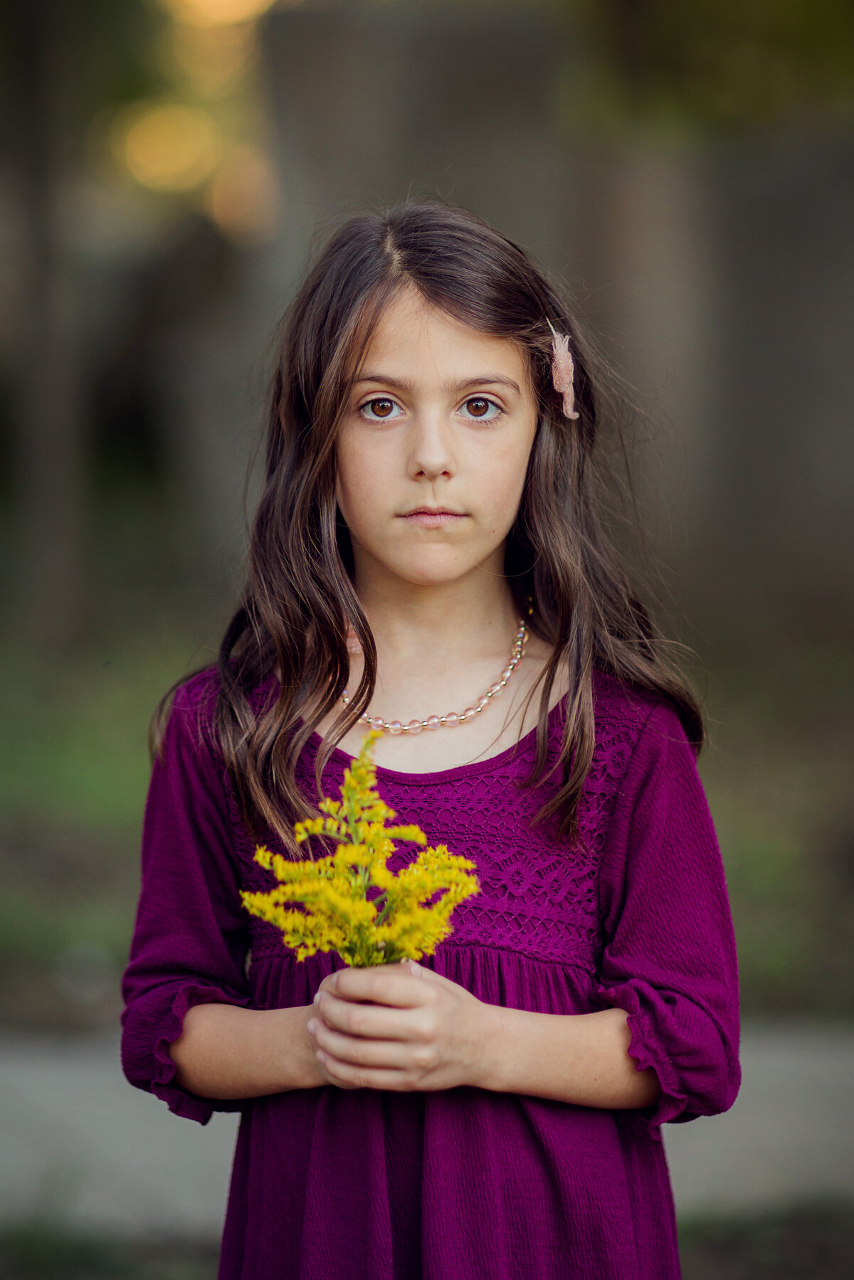 A young girl in a pink dress is looking at the camera, holding yellow flowers.