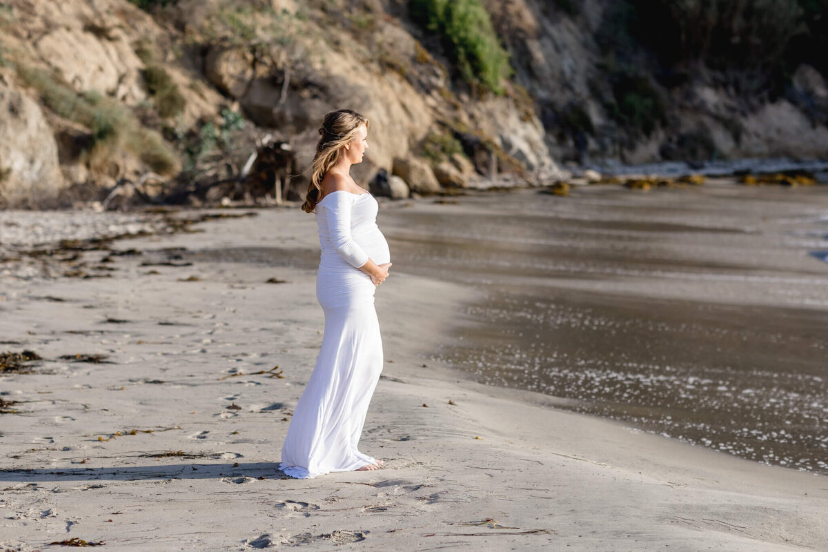 A woman in a long, white dress stands on a sandy beach, gazing at the ocean. Her hands rest gently on her pregnant belly. The shoreline is lined with rocks and greenery in the background.