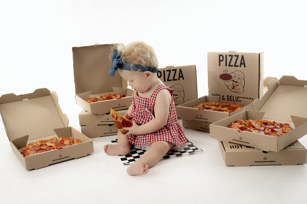 A toddler girl looks down at a slice of pizza in her hand