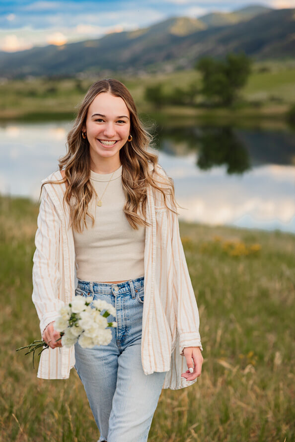 Girl walks towards camera with flowers in her hand and a lake behind her.