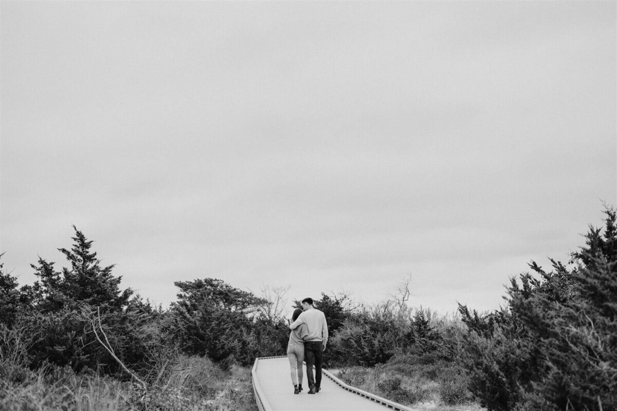 The engaged couple walking while hugging, on a wooden bridge amidst grasses and plants in New York. Engagement Image by Jenny Fu Studio