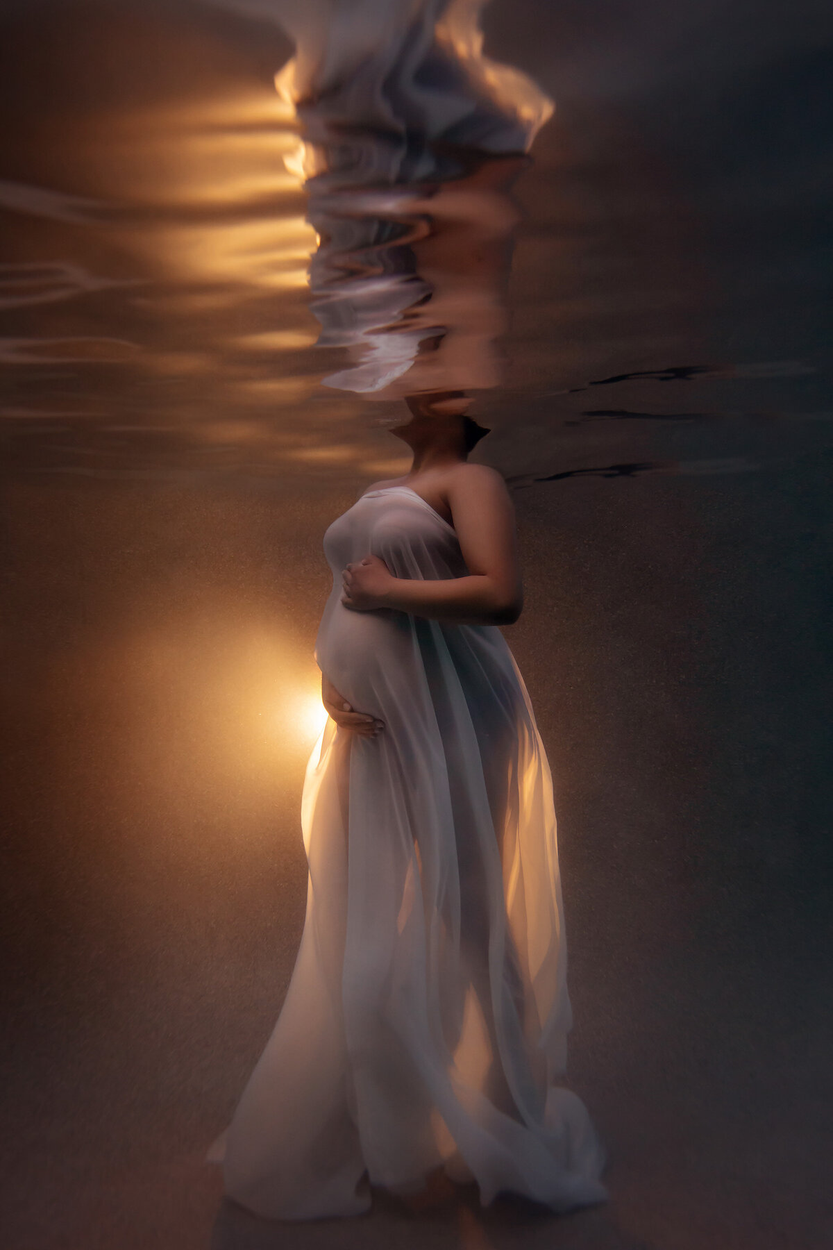 A woman standing gracefully underwater, draped in a flowing sheer white gown illuminated by warm golden light. Her reflection ripples above, creating a dreamy, abstract effect. The serene atmosphere emphasizes elegance and tranquility.