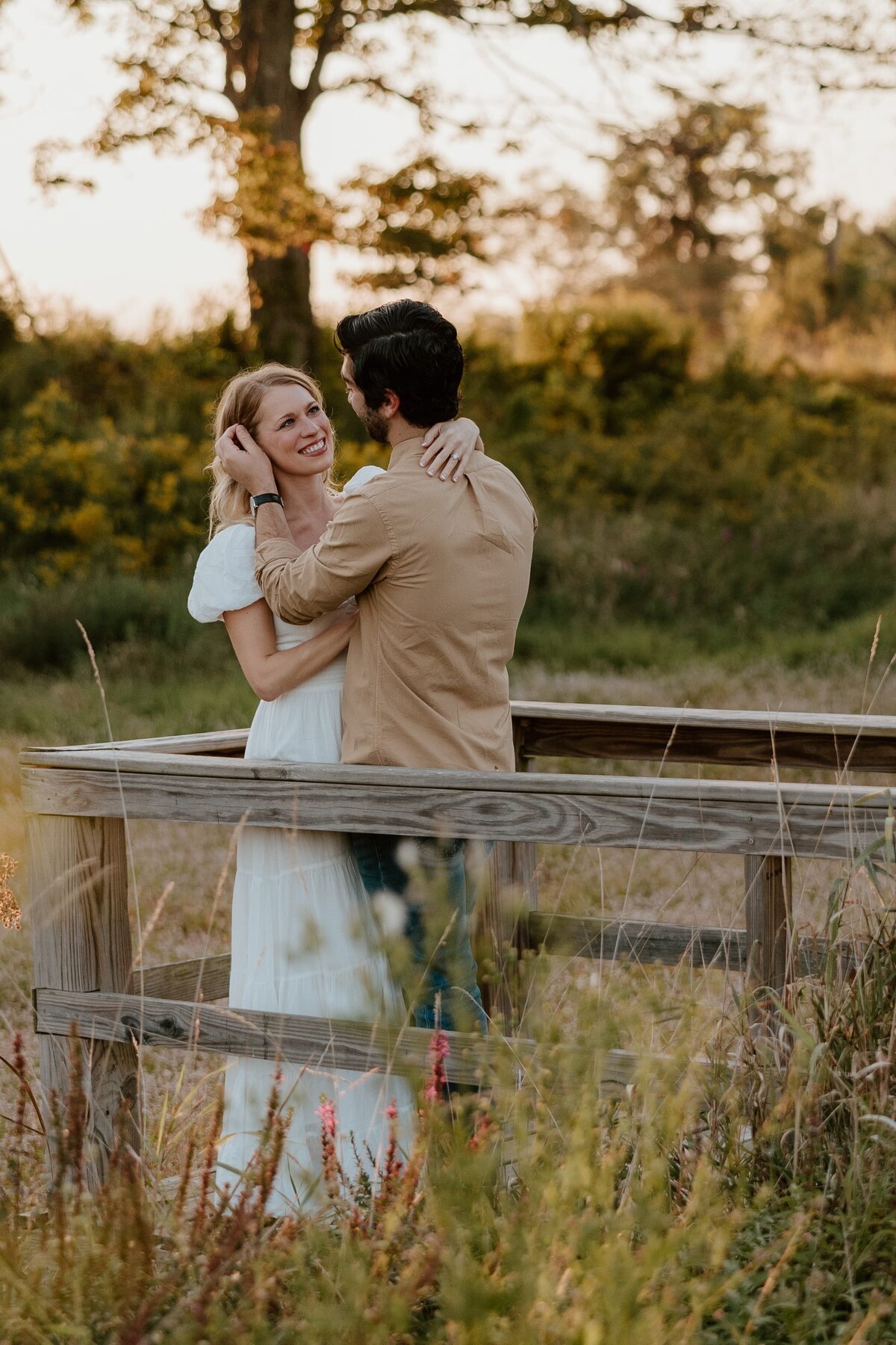 A couple embracing on a rustic wooden platform surrounded by tall grass and wildflowers during golden hour. The woman wears a flowing white dress, and the man wears a tan shirt and jeans. They are gazing at each other with smiles, with a backdrop of trees and soft evening light.