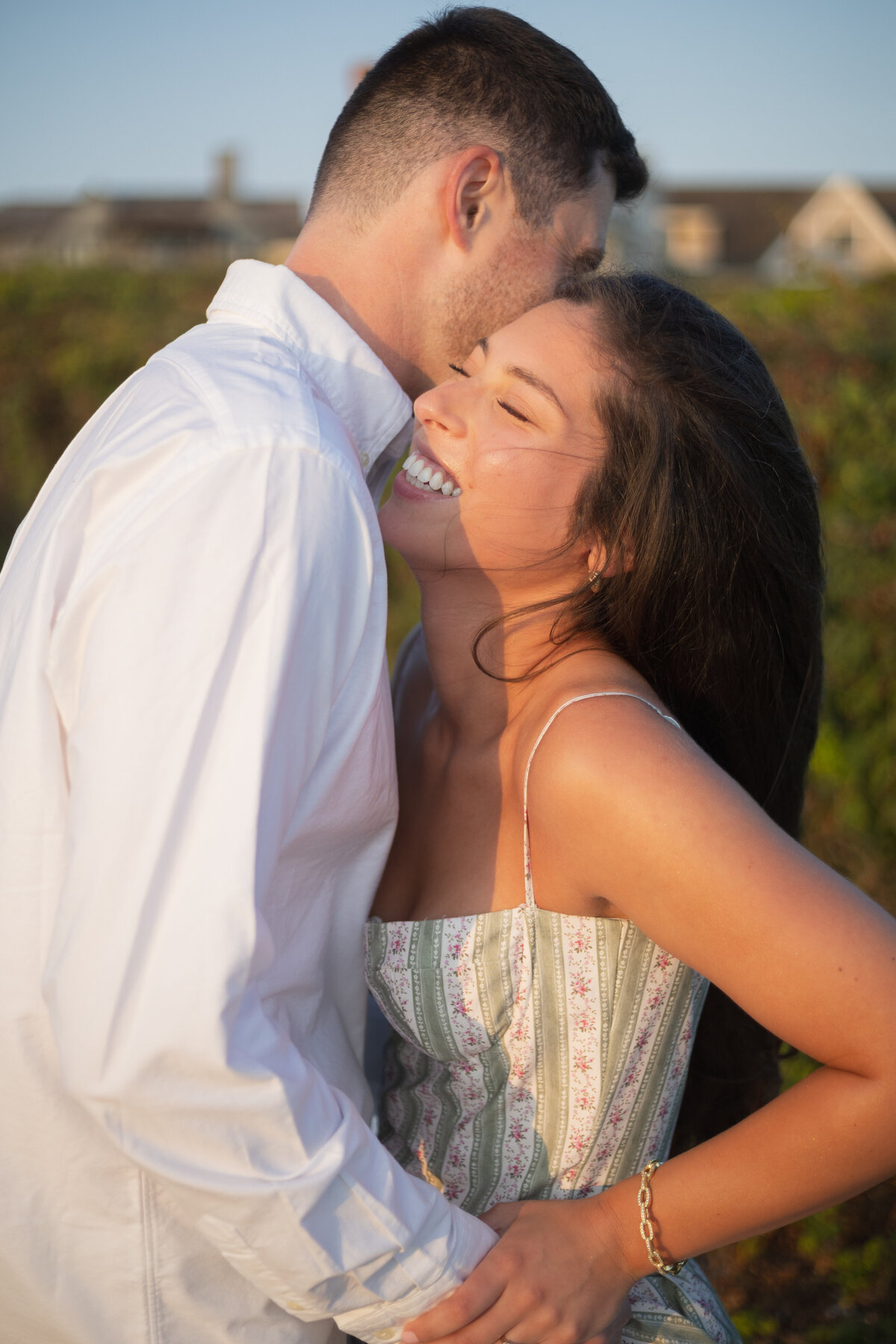 Collins Beach Newport RI | Kelsey Sheehan Photography Timeless Rhode Island Weddings | A woman in a sundress laughing joyfully, leaning close to a man in a white shirt. They are outdoors on a sunny day, with greenery in the background.