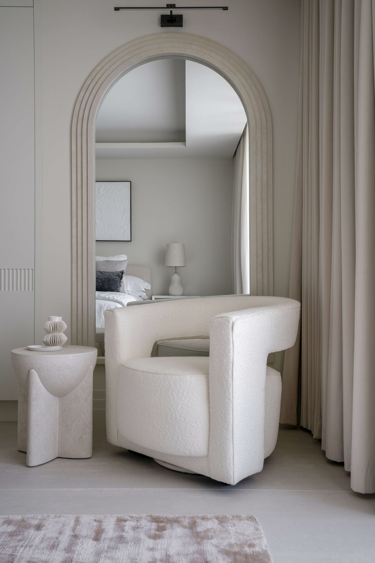 Contemporary white armchair in the corner of a light and airy guest bedroom. Behind the chair is a large arched mirror with a neutral frame.