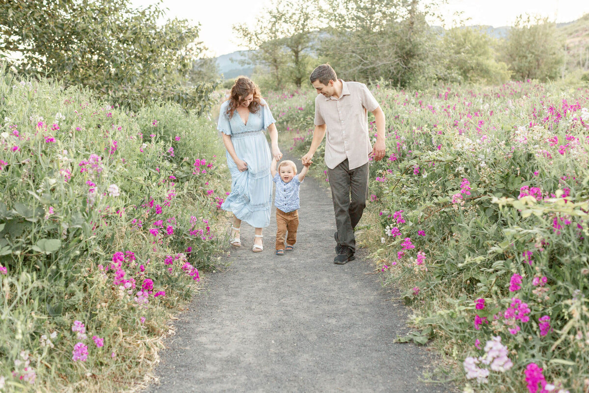 Family of three, Mom, toddler boy and dad walking hand in hand down a path that is surrounded by tall pink and white wildflowers. There are mountains off to the distance behind them as the sun is beginning to get lower in  the sky.