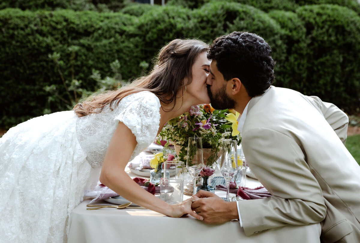Bride and groom sharing a quiet moment at a table set up in a garden, surrounded by spring greenery.