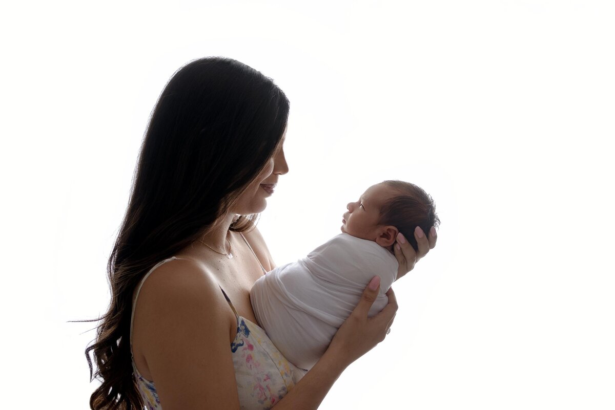 Profile backlit image of mom holding newborn baby out and looking down at him