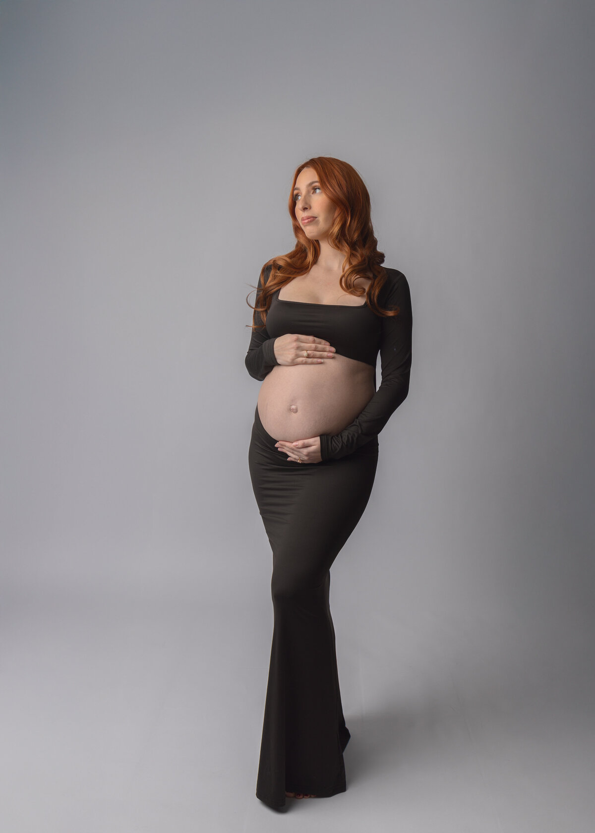 Elegant maternity portrait shows an expectant mother in a fitted black dress against a neutral gray backdrop.