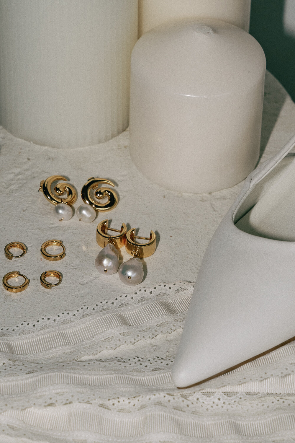 Flat lay of gold hoop earrings, pearls, and a white clutch bag on neutral linen with large white candles