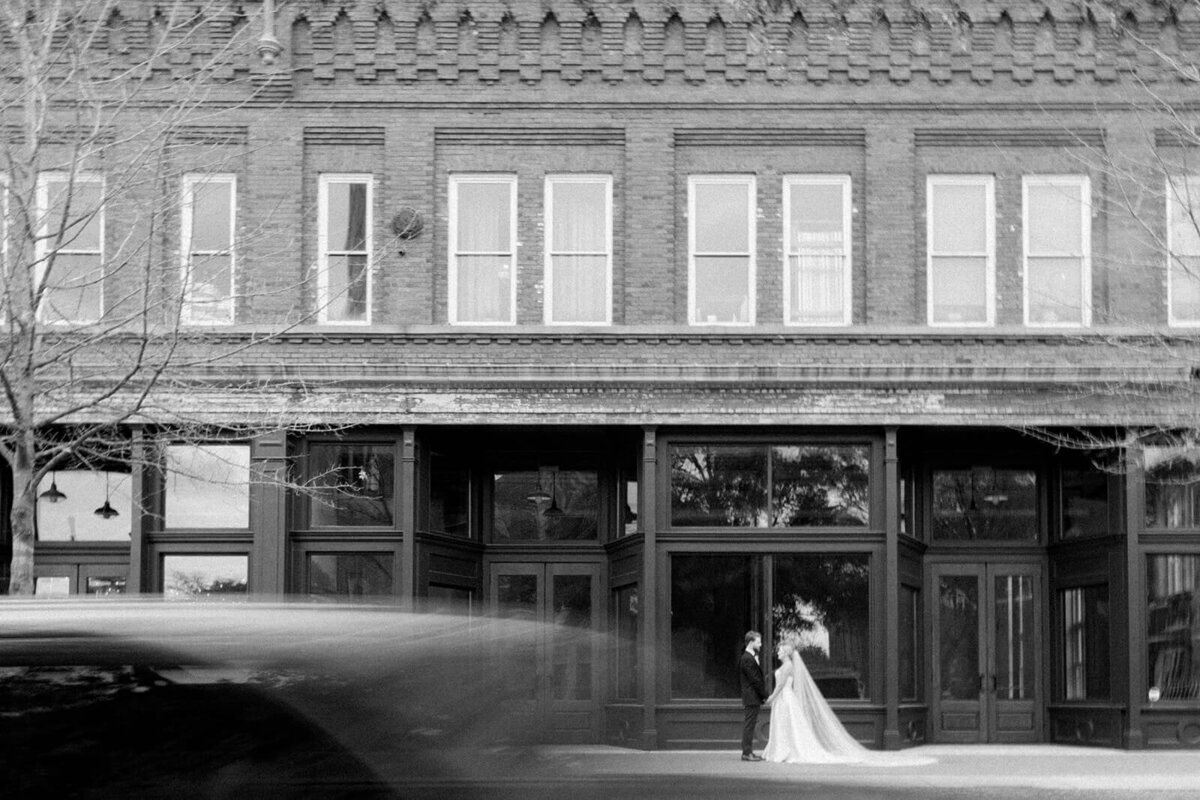 Black and white wedding photo of a bride and groom standing in front of a historic brick building in South Carolina. The motion blur of a passing car adds a dynamic, documentary-style feel to this timeless and artistic wedding portrait. Captured by a Columbia, SC wedding photographer specializing in authentic and cinematic wedding imagery.