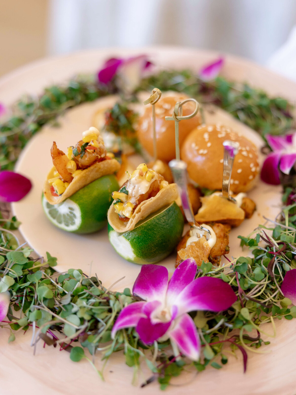 A decorative platter features small sandwiches and tacos nestled in lime halves, surrounded by fresh greens and adorned with vibrant purple flowers.