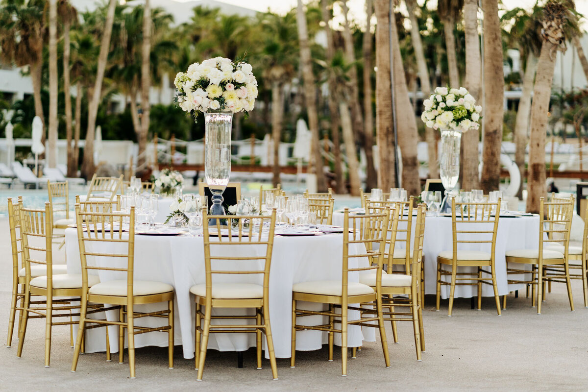 Outdoor reception tables with gold chairs and tall floral centerpieces, palm trees in the background.