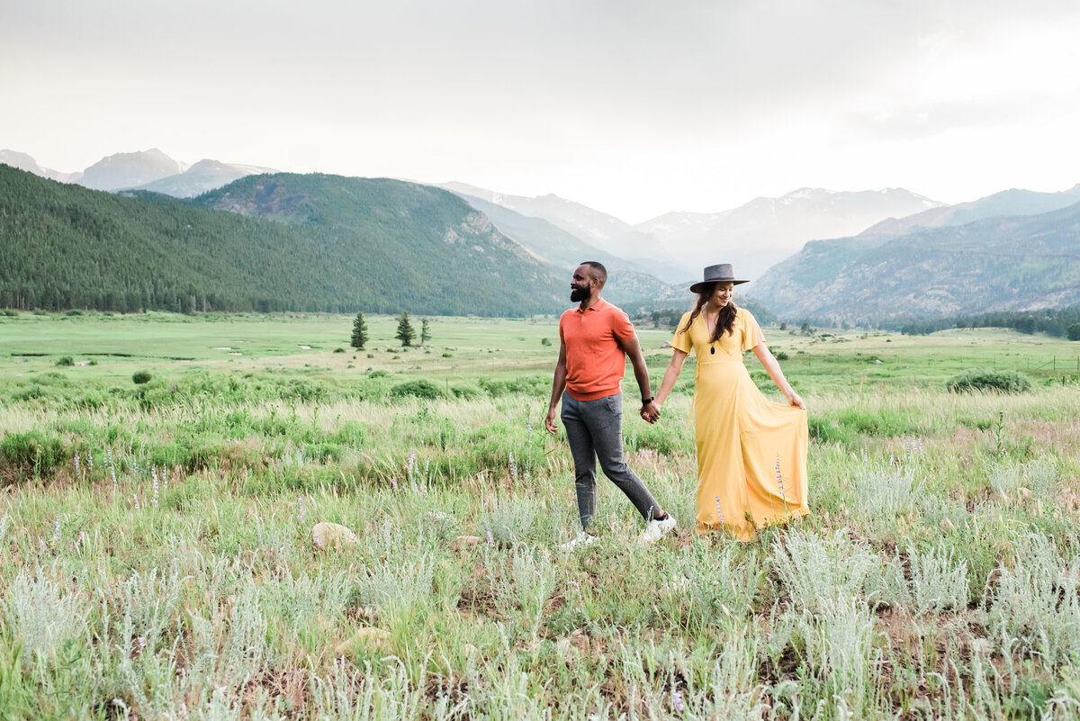 Denver wedding photographer captures a couple during their engagement photos at Rocky Mountain National Park walking through a field with mountains in the background