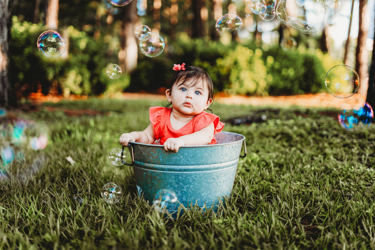 Baby girl sitting in a metal bucket surrounded by bubbles during a playful outdoor portrait session in Orlando, Florida