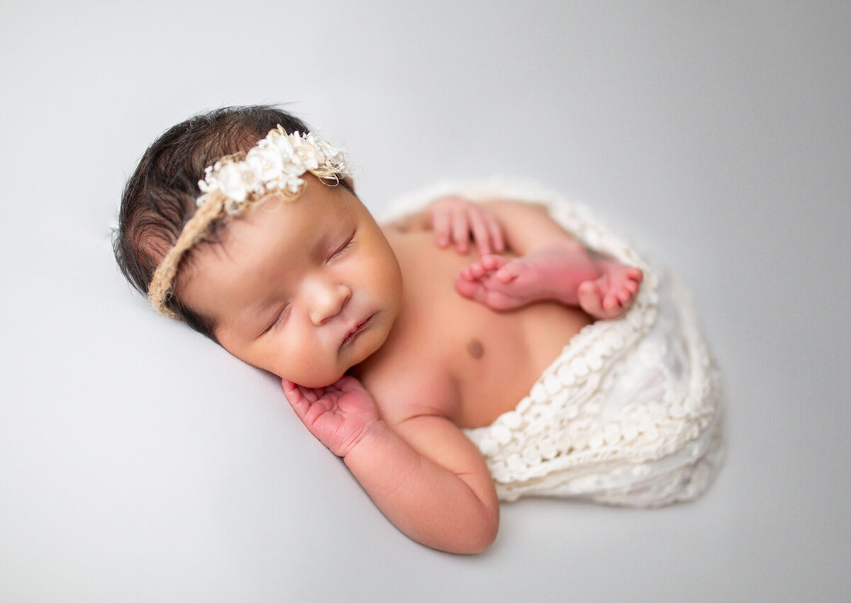 Baby girl on white backdrop with hand resting under cheek and wrapped in lace 
