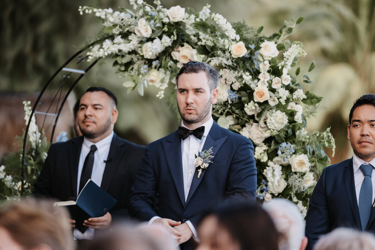 Groom looking down the aisle seeing bride  with florals in the backgroudn