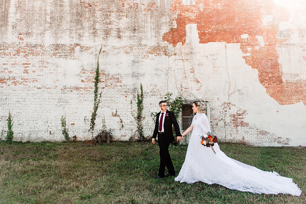 The bride and groom walk hand in hand  in a grassy field next to a white washed wall streaked with  red. The groom is wearing a black suit with a white shirt and red tie. The bride is wearing a white dress with a cathedral length train. she is holding a bouquet of red and white flowers down at her side as she holds hands with the groom.