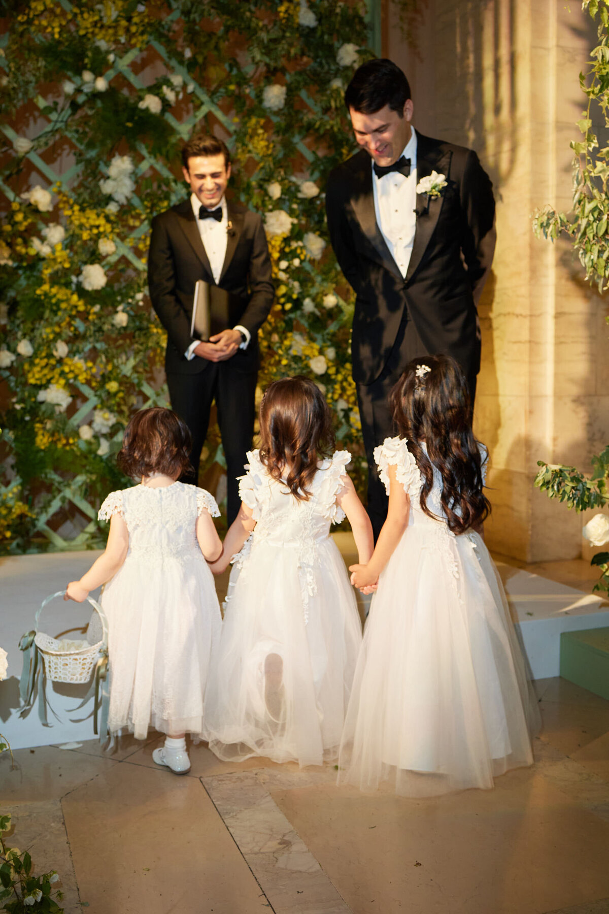Three young flower girls in white dresses walk hand-in-hand down the aisle towards a smiling groom and officiant at a wedding ceremony. The background features greenery and floral decorations, creating a charming outdoor ambiance. This lovely moment was captured at the New York Public Library in New York City by photographer Shawn Connell. Videographers Aaron Navak Films were also present to beautifully document this beautiful wedding ceremony.