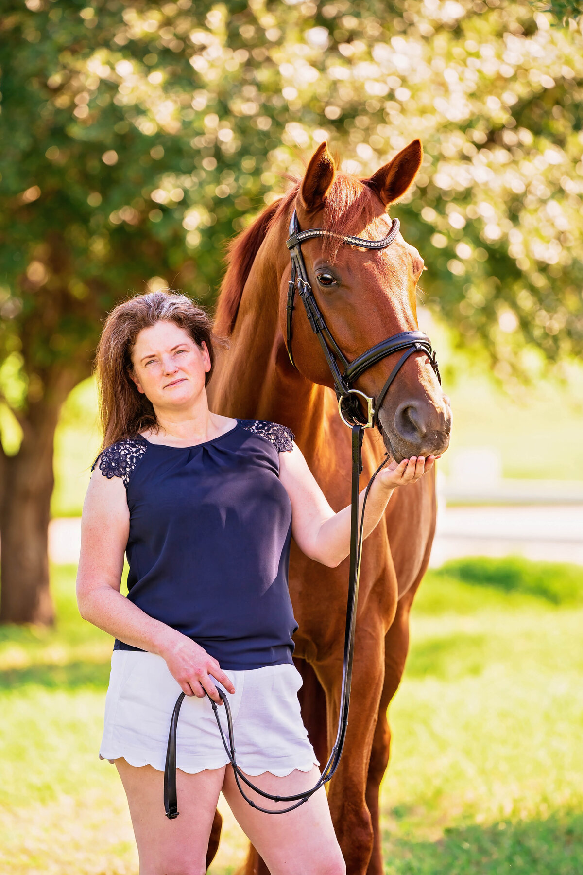 Female equestrian poses in shorts and a blue shirt while her chestnut warmblood horse looks over her shoulder and nuzzles her hand.