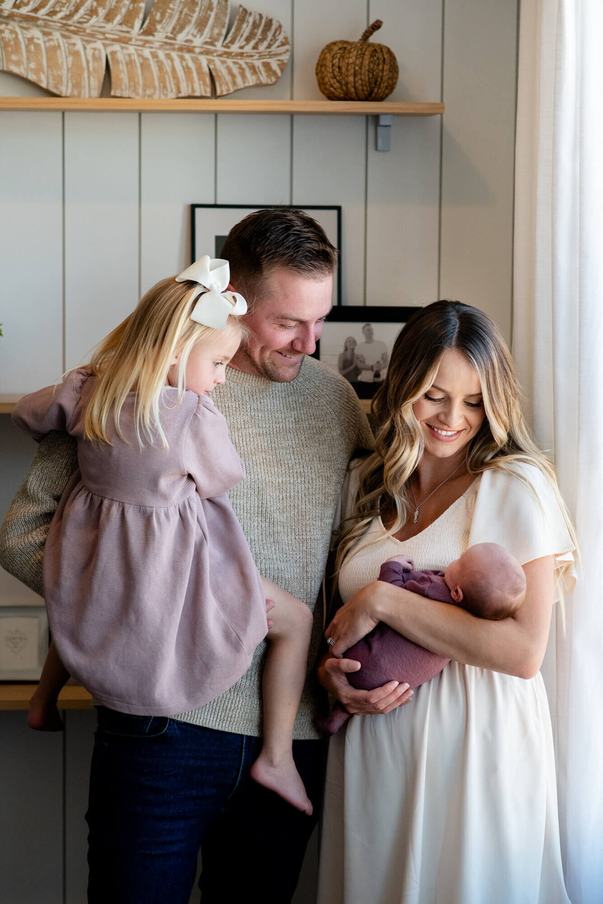 A mom and dad stand in a window smiling with their newborn baby and toddler daughter in their arms