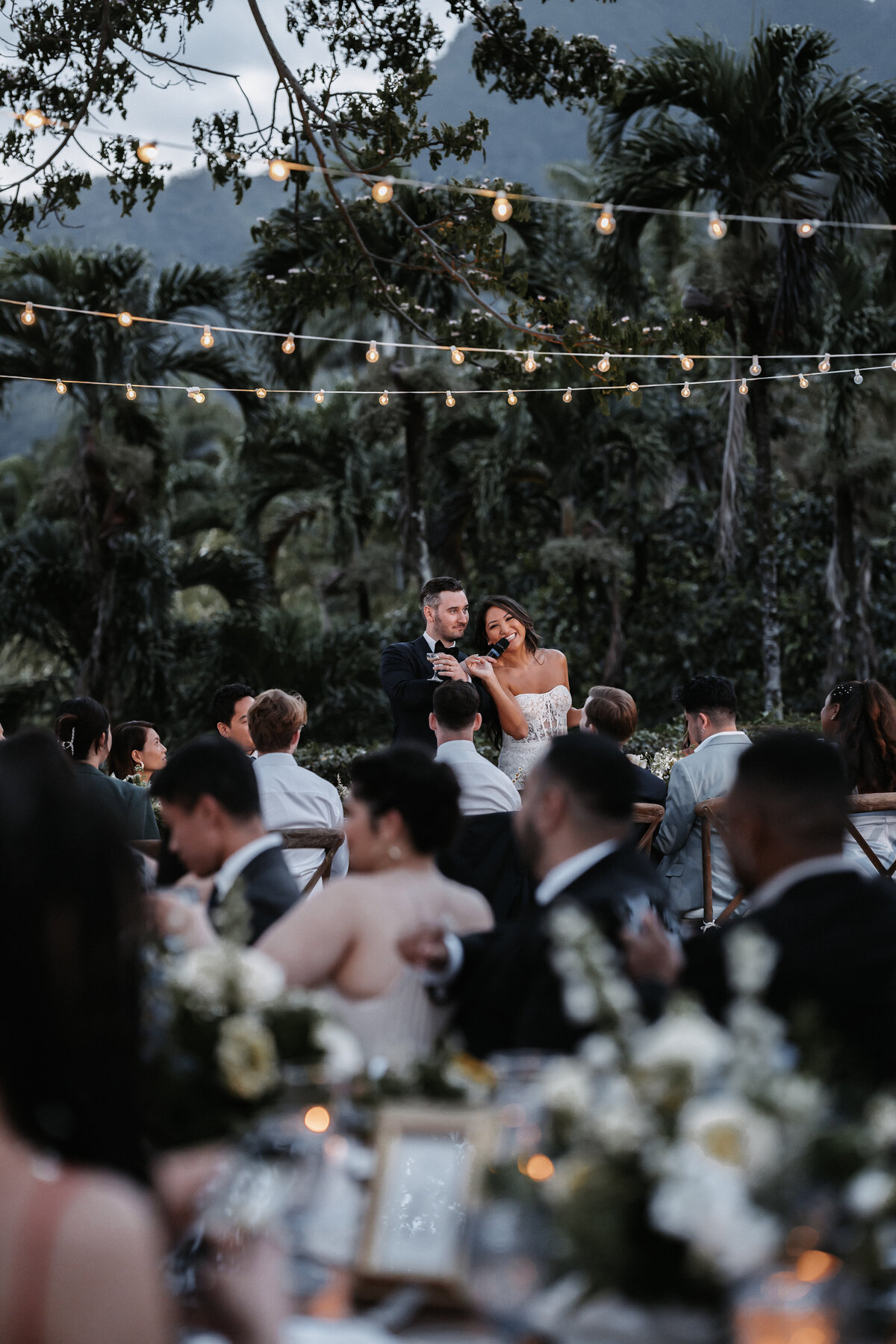 Bride and groom standing at dusk holding a microphone speaking to their guests