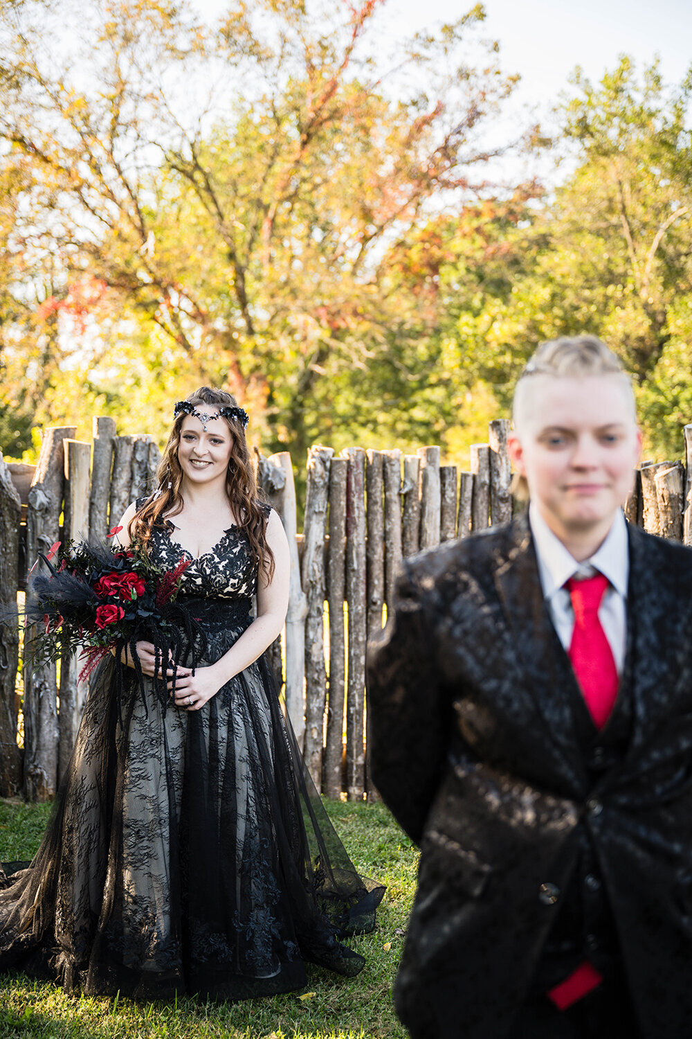 Two LGBTQ+ brides stand in the backyard of an Airbnb they rented in Roanoke, Virginia on their elopement. One bride, dressed in a black floral suit, stands in front of another bride, who is wearing a black dress. The bride in the foreground is purposefully out of focus to focus on the bride in the background who is smiling as they wait for their first look moment.