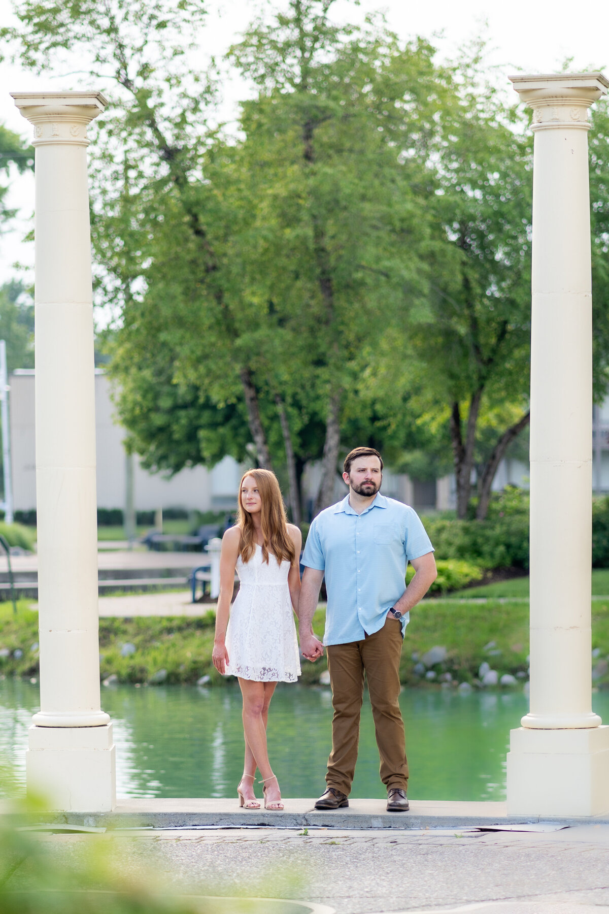 engaged couple holding hands at a park in Grosse Pointe Michigan between two white pillars looking off opposite directions into the distance