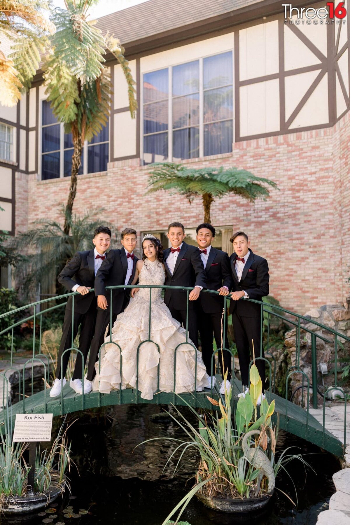Young men dressed in black tuxedos pose with the quinceanera girl on a small bridge