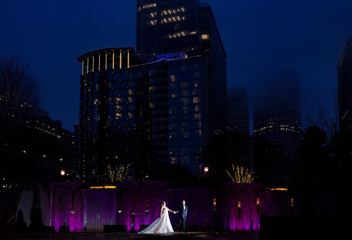 Groom leading the bride in a nighttime portrait by the water feature at Romare Bearden Park. The water feature glows purple, with the city lights twinkling in the background beneath a deep blue sky.
