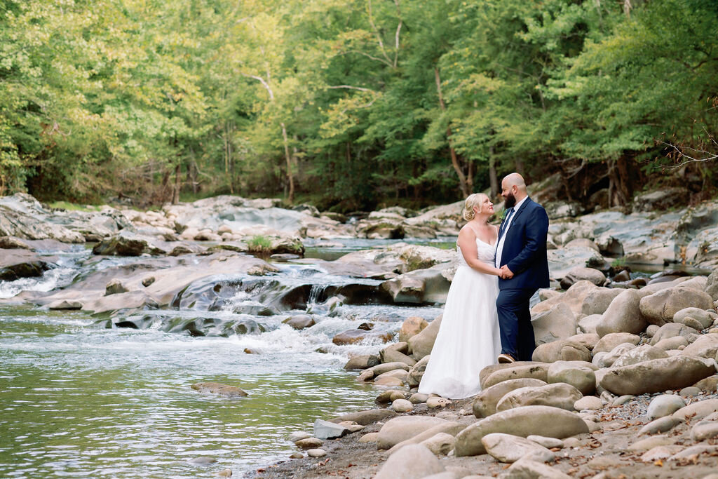 Bride and groom standing on a rocky riverbank at Greenbrier, holding hands and smiling at each other during their eloping to Gatlinburg ceremony, surrounded by flowing water and lush green forest.
