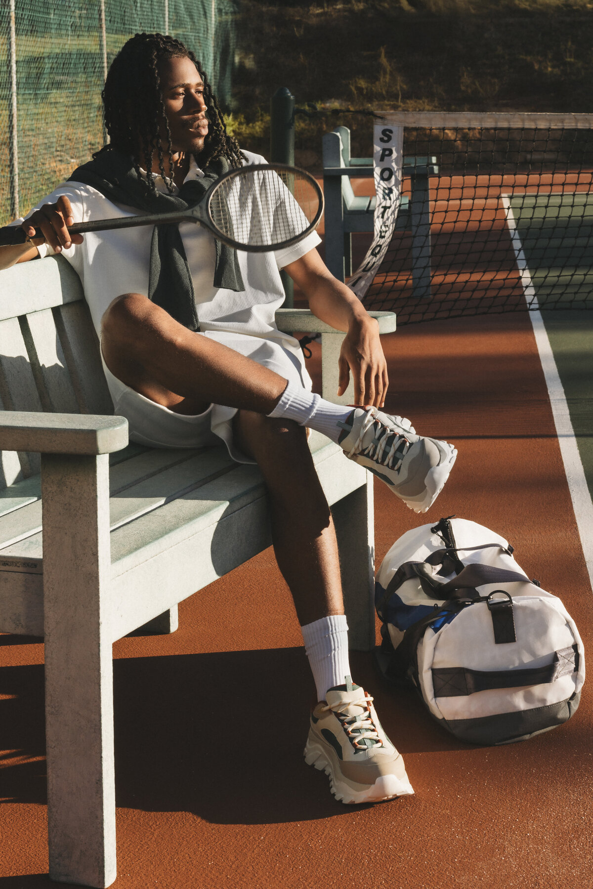 Man relaxing on a tennis bench at sunset, holding a vintage racket with a Swim Essentials duffel bag by his side, dressed in sporty casualwear.