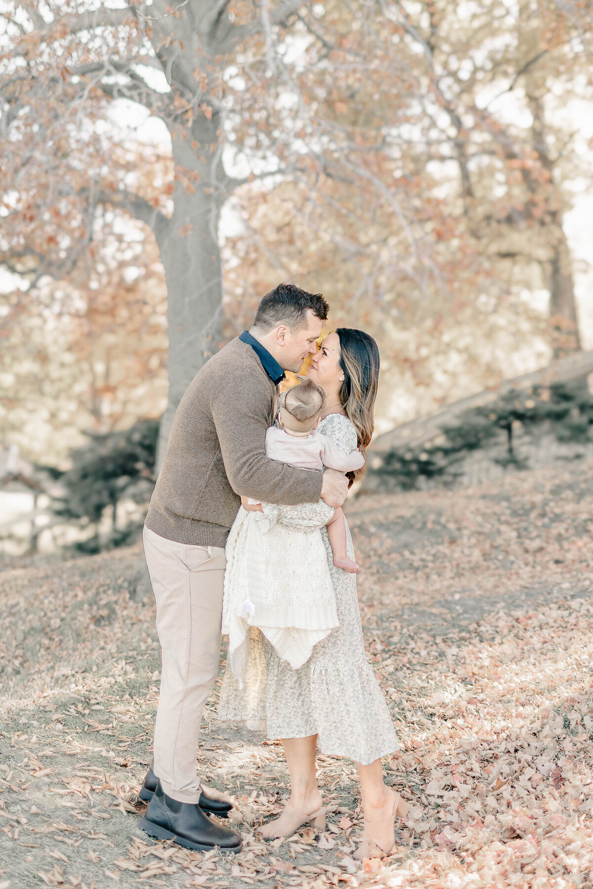 Parents embracing each other while holding their baby girl in a sunlit park during her Kansas City birthday milestone session.