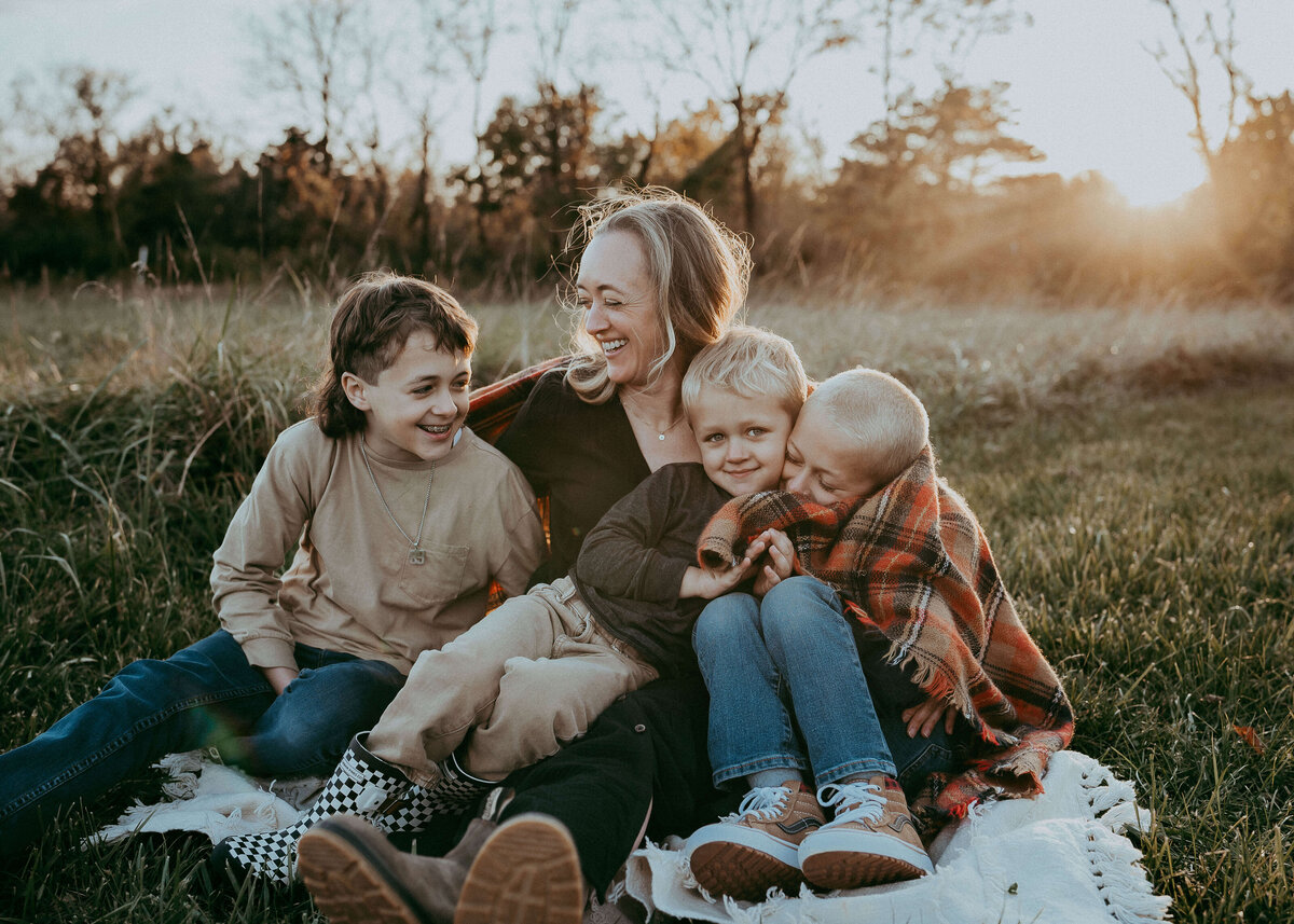 A mom laughs with her three wild boys while sitting on a blanket on their family farm being back, let by a gorgeous sunset and autumn colors.
