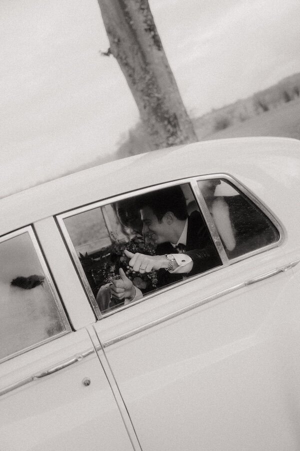 Marié, pouces levés, souriant à travers la vitre d'une voiture ancienne. Photo par Kamescop, photo et vidéo de mariage en Bretagne.