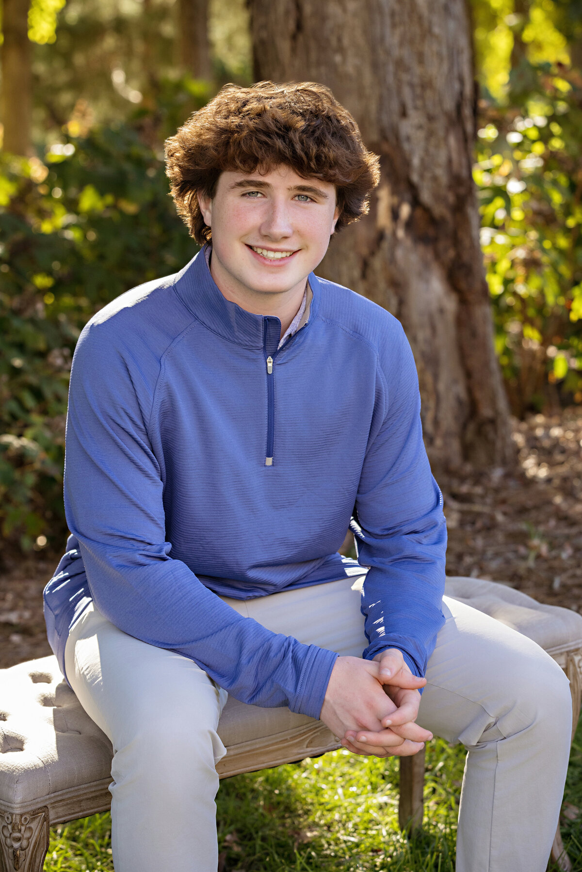 Confident portrait of a high school teen son seated on a garden bench framed by greenery in Ballantyne Charlotte NC, wearing a blue quarter‑zip sweater over a button‑down