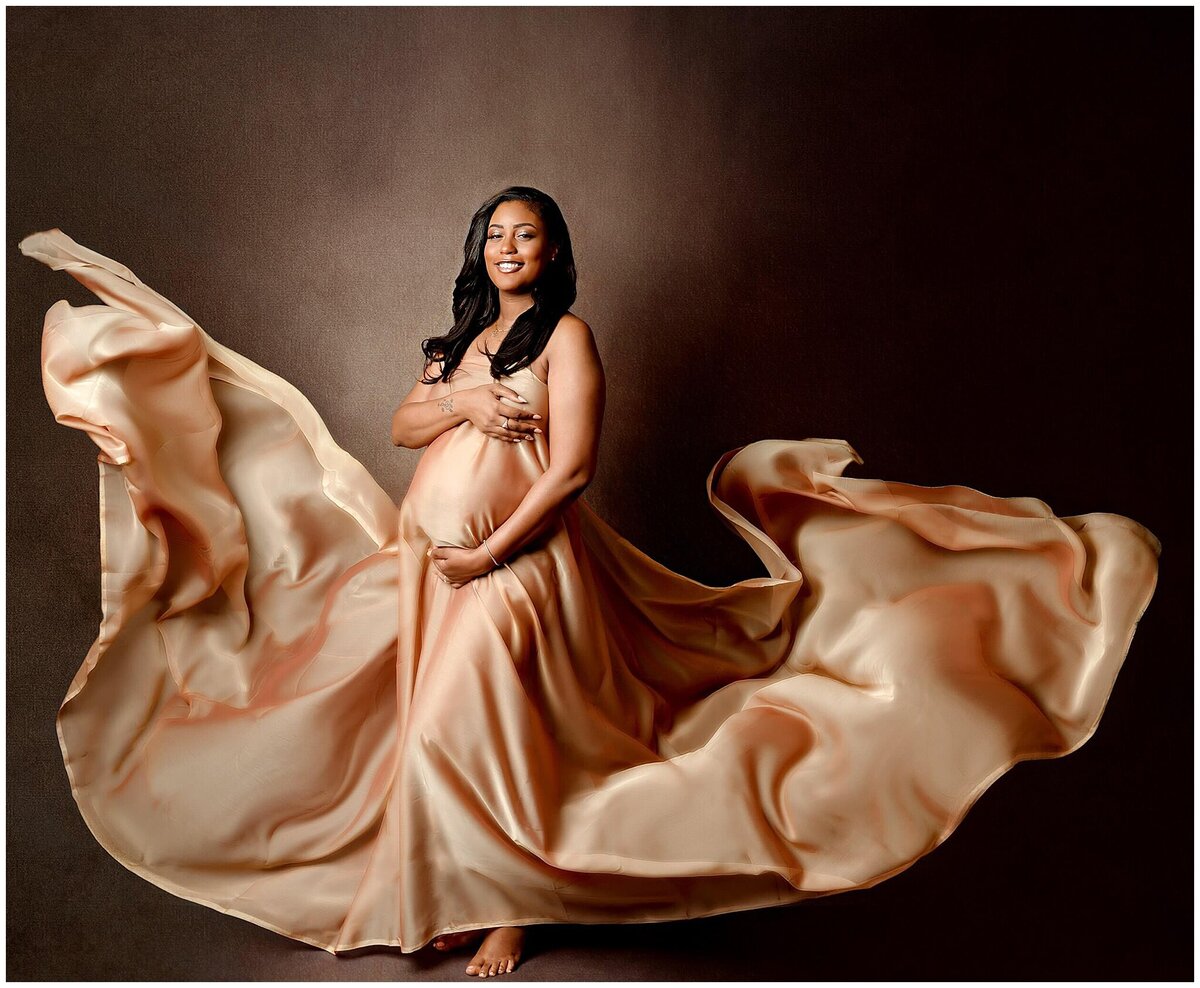 In a maternity studio session a pregnant woman stands gracefully as a vibrant gold fabric is tossed around her. The flowing fabric adds a sense of movement and elegance to the image.